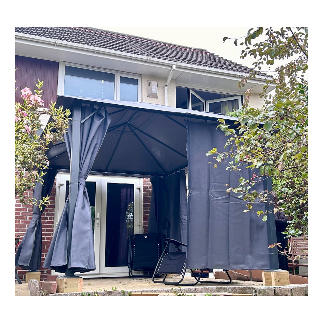 A gazebo with grey curtains in a garden with rose bushes leading from the house.