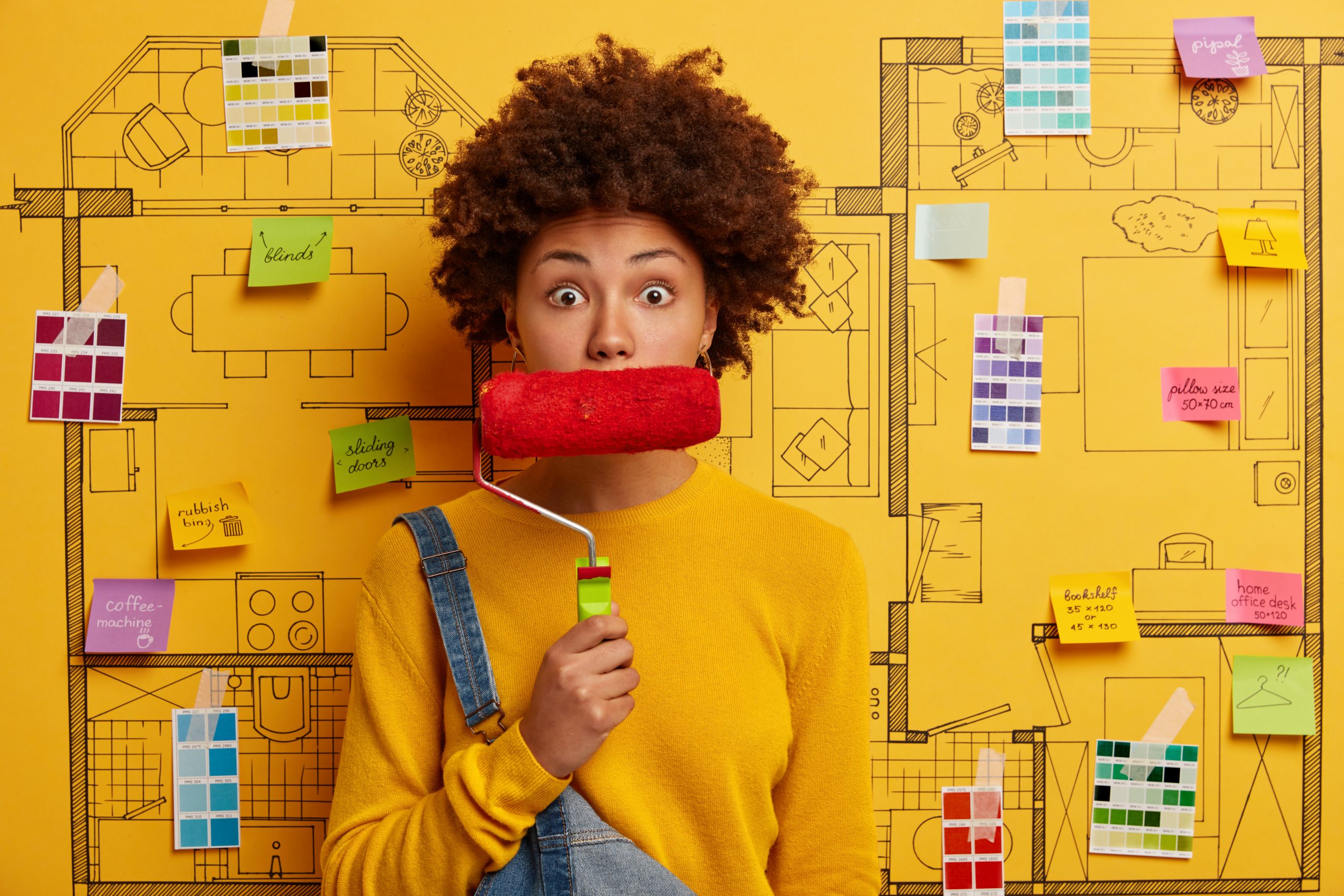 Shocked emotional woman has curly hairstyle keeps paint roller on mouth, stares with bugged eyes, surprised to have much work to do, stands against home interior design. Room plan with drawn furniture